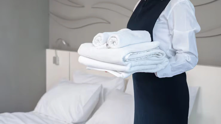 Lodging & Travel Linen Services 4 servicio habitacion hotel haciendo habitacion mujer joven camarera sosteniendo pila toallas bano blancas dobladas pie junto cama habitacion invitados 63313 1772
